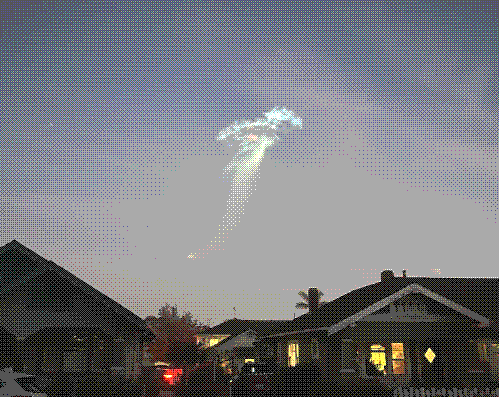 A glowing jellyfish-shaped cloud at dusk. Taken on a residential street, there are two houses in the bottom quarter of the image, with soft yellow light diffused through the windows.